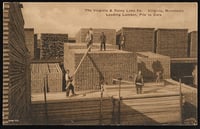 Stacks of lumber rise to the sky in stacks on two different levels above the ground, with workers posed on on each of them.